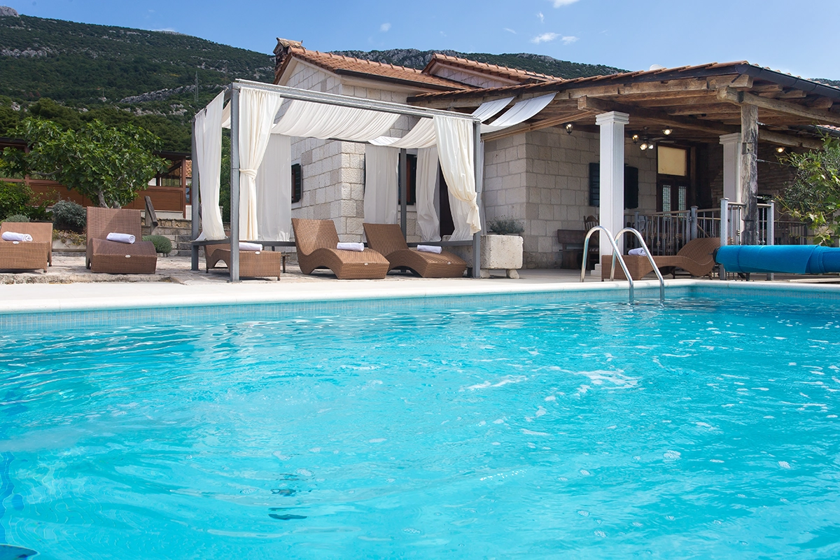 Pool and sun loungers with the front side of the villa during the day.